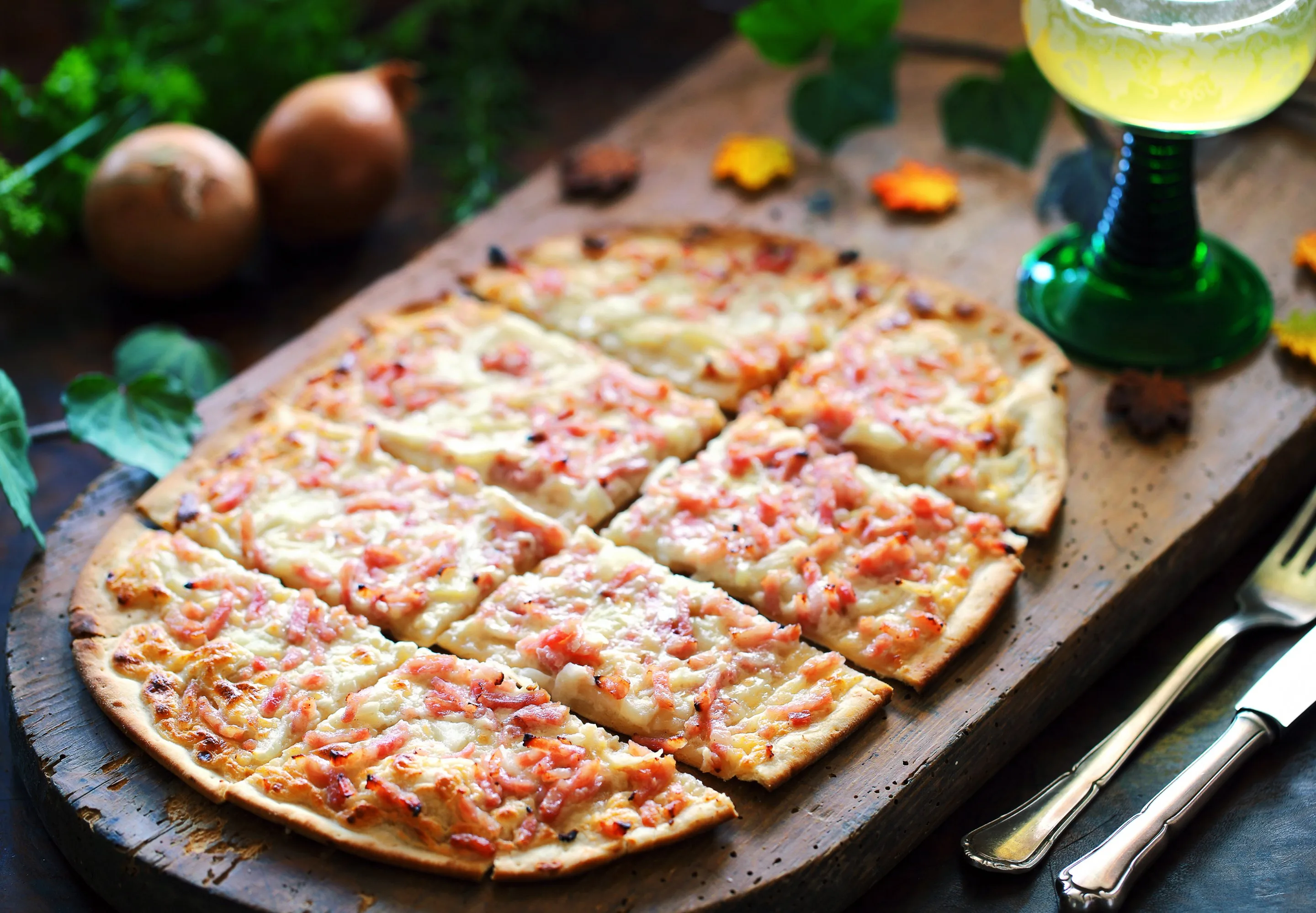 Flammkuchen mit Speck und Zwiebeln, in Stücke geschnitten, auf einem Holzbrett mit Besteck und einem Glas.
